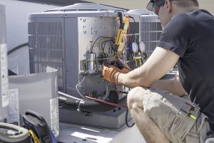 Air conditioner repair in Mantoloking being done on condenser by mand in cargo shorts, black t-shirt, safety goggles, work gloves.
